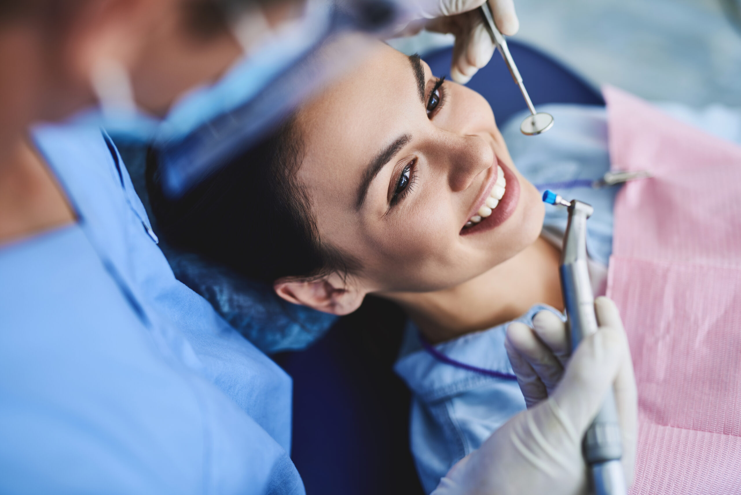 Young lady receiving teeth cleaning and polishing at clinic Close up portrait of charming woman sitting in dental chair while stomatologist holding polisher and mirror. Girl is smiling